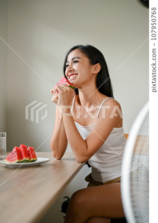 A smiling Asian woman enjoys her fresh watermelons while sitting in front of a fan 107950768