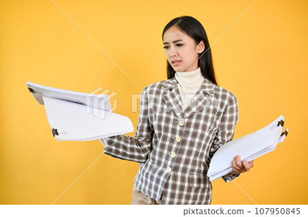 A perplexed Asian businesswoman stands against an isolated yellow background with documents A perplexed Asian businesswoman stands against an isolated yellow background with documents 107950845