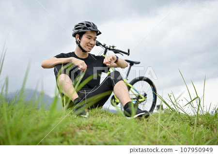 A happy Asian man checks his calories burned on his smartwatch while resting on the grass A happy Asian man checks his calories burned on his smartwatch while resting on the grass 107950949