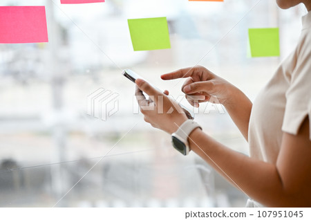Close-up image of a woman using her smartphone while standing by the window in her office. Close-up image of a woman using her smartphone while standing by the window in her office. 107951045