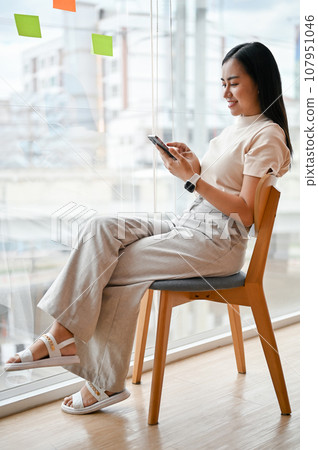 An attractive Asian woman sits at a chair by the window and uses her smartphone. 107951046