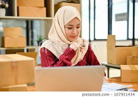 A focused young Asian Muslim female online seller checking products stocks A focused young Asian Muslim female online seller checking products stocks 107951434