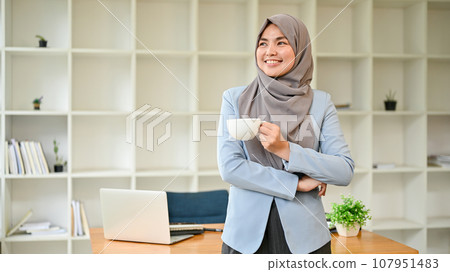 A confident Asian Muslim businesswoman stands in her office with a coffee cup in her hands. 107951483