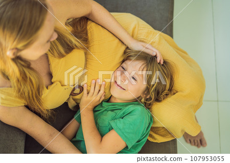 A heartwarming moment: pregnant mother and her older son sit on the sofa, engaging in a tender conversation about the upcoming pregnancy A heartwarming moment: pregnant mother and her older son sit on the sofa, engaging in a tender conversation about the upcoming pregnancy 107953055