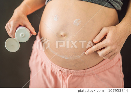 Positive young pregnant woman in comfortable homewear applying belly butter on her big tummy, woman belly Positive young pregnant woman in comfortable homewear applying belly butter on her big tummy, woman belly 107953844