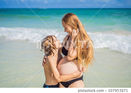A picturesque moment: a pregnant woman and her son enjoying the turquoise sea, a heartwarming family bond by the tranquil shore 107953871