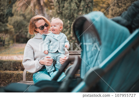 Mother sitting on bench in urban park, laughing cheerfully, holding her smiling infant baby boy child in her lap having baby stroller parked by their site. 107955727