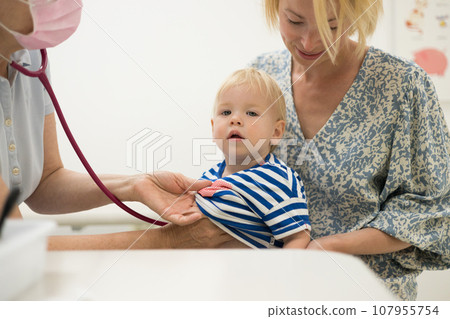 Infant baby boy child being examined by his pediatrician doctor during a standard medical checkup in presence and comfort of his mother. National public health and childs care care koncept Infant baby boy child being examined by his pediatrician doctor during a standard medical checkup in presence and comfort of his mother. National public health and childs care care koncept 107955754