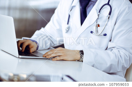 Unknown male doctor sitting and working with laptop in clinic at his working place, close-up. Young physician at work. Perfect medical service, medicine concept Unknown male doctor sitting and working with laptop in clinic at his working place, close-up. Young physician at work. Perfect medical service, medicine concept 107959033