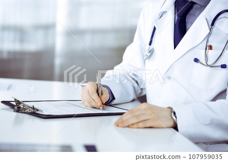 Unknown male doctor sitting and working with clipboard of medication history record in clinic at his working place, close-up. Young physician at work. Perfect medical service, medicine concept Unknown male doctor sitting and working with clipboard of medication history record in clinic at his working place, close-up. Young physician at work. Perfect medical service, medicine concept 107959035