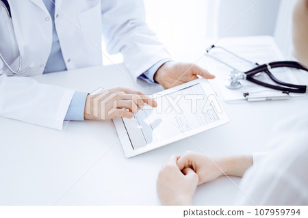 Doctor and patient sitting opposite each other at the desk in clinic. The focus is on female physician's hands pointing into tablet computer touchpad, close up. Medicine concept Doctor and patient sitting opposite each other at the desk in clinic. The focus is on female physician's hands pointing into tablet computer touchpad, close up. Medicine concept 107959794
