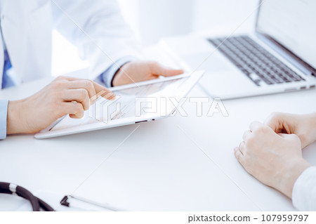 Doctor and patient sitting opposite each other at the desk in clinic. The focus is on female physician's hands using tablet computer, close up. Medicine concept Doctor and patient sitting opposite each other at the desk in clinic. The focus is on female physician's hands using tablet computer, close up. Medicine concept 107959797