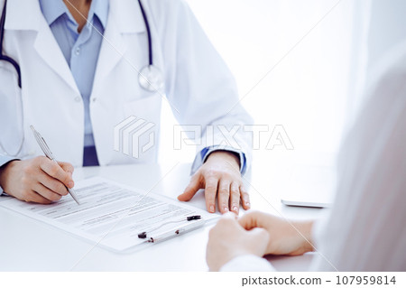 Doctor and patient sitting opposite each other at the desk in clinic. The focus is on female physician's hands filling up the medication history record form or checklist, close up. Medicine concept 107959814