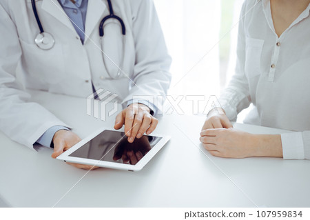 Doctor and patient sitting near each other at the desk in clinic. The focus is on female physician's hands pointing into tablet computer touchpad, close up. Medicine concept Doctor and patient sitting near each other at the desk in clinic. The focus is on female physician's hands pointing into tablet computer touchpad, close up. Medicine concept 107959834