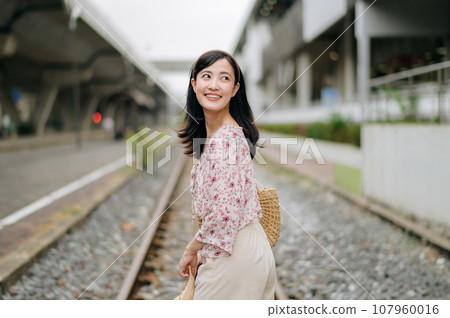 young asian woman traveler with weaving basket happy smiling looking to a camera beside train railway. Journey trip lifestyle, world travel explorer or Asia summer tourism concept. 107960016