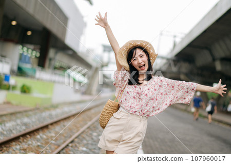 young asian woman traveler with weaving basket happy smiling looking to a camera beside train railway. Journey trip lifestyle, world travel explorer or Asia summer tourism concept. 107960017