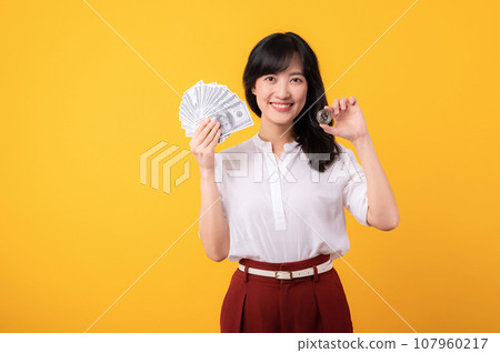 Portrait beautiful young asian woman enterpriser happy smile wearing white shirt and red plants holding cash dollar money and crypto digital currency isolation on yellow background. Wealth concept. Portrait beautiful young asian woman enterpriser happy smile wearing white shirt and red plants holding cash dollar money and crypto digital currency isolation on yellow background. Wealth concept. 107960217