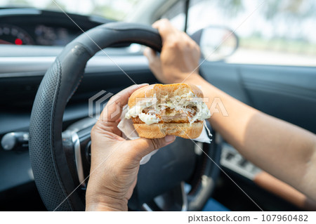 Asian lady holding hamburger to eat in car, dangerous and risk an accident. Asian lady holding hamburger to eat in car, dangerous and risk an accident. 107960482