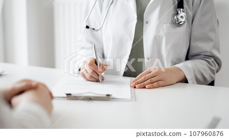 Doctor and patient sitting near each other at the white desk in clinic. Female physician is listening filling up a records form. Medicine concept Doctor and patient sitting near each other at the white desk in clinic. Female physician is listening filling up a records form. Medicine concept 107960876