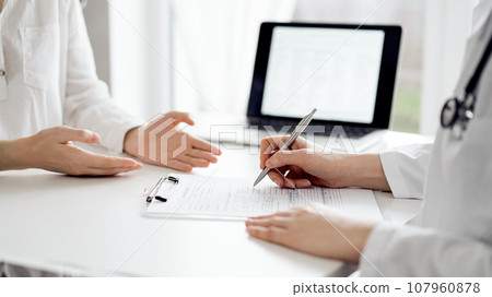 Doctor and patient sitting and discussing something near each other at the white desk in clinic. Female physician is listening filling up a records form. Medicine concept Doctor and patient sitting and discussing something near each other at the white desk in clinic. Female physician is listening filling up a records form. Medicine concept 107960878