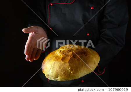 The baker presents freshly baked wheat bread in his hand. Place for advertising on a black background. The concept of baking round bread The baker presents freshly baked wheat bread in his hand. Place for advertising on a black background. The concept of baking round bread 107960919