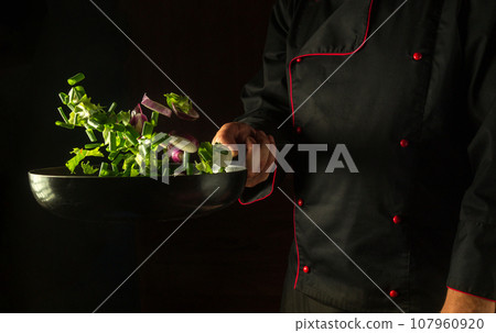 The cook prepares fresh vegetables in a frying pan. Grande cuisine on dark background. Free advertising space. Frying pan with flying vegetables in the chef hand. 107960920