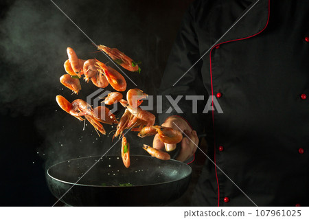 The chef cooks shrimp in a hot pan with steam. The concept of cooking seafood or healthy vegetarian food and eating on a black background. Freeze in motion. Free space for recipe 107961025