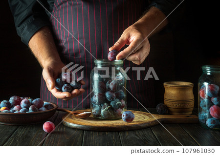 Filling a jar with plums. Preparing a sweet drink from fresh plum and sugar on the kitchen table. The process of preserving berries in a jar 107961030