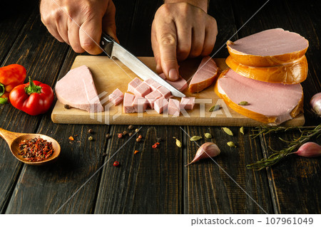 Close-up of a cook hands cutting scalded sausage into small pieces with a knife to prepare meat dishes for lunch. Copy space Close-up of a cook hands cutting scalded sausage into small pieces with a knife to prepare meat dishes for lunch. Copy space 107961049