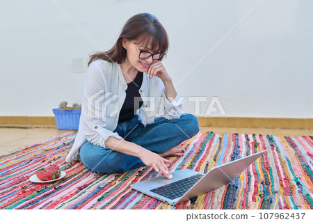 Happy middle-aged woman sitting on floor at home using laptop Happy middle-aged woman sitting on floor at home using laptop 107962437