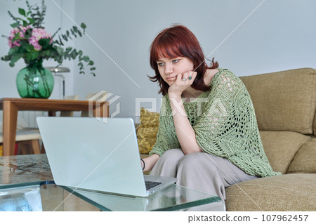 Young female sitting on couch at home using laptop computer Young female sitting on couch at home using laptop computer 107962457