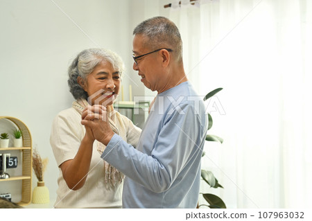 Shot of romantic elderly couple spouses dancing in living room and looks at each other with love 107963032