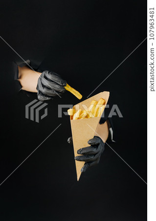 Hand holding French fries through torn holes in black paper background. Street food concept 107963481