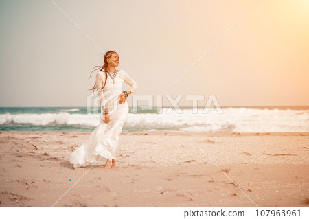 woman sea white dress. Model in boho style in a white long dress and silver jewelry on the beach. Her hair is braided, and there are many bracelets on her arms. 107963961