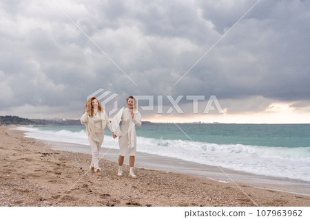 Women sea walk friendship spring. Two girlfriends, redhead and blonde, middle-aged walk along the sandy beach of the sea, dressed in white clothes. Against the backdrop of a cloudy sky and the winter Women sea walk friendship spring. Two girlfriends, redhead and blonde, middle-aged walk along the sandy beach of the sea, dressed in white clothes. Against the backdrop of a cloudy sky and the winter 107963962