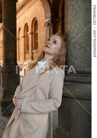 Beautiful young woman with red lips in beige coat stands near old building and thinking about something. 107964062