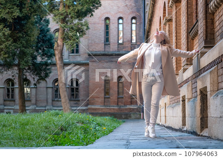 Happy young woman in white clothes near old red brick building looking up. Attractive Girl walks alone. Happy young woman in white clothes near old red brick building looking up. Attractive Girl walks alone. 107964063
