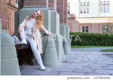 Woman waiting someone and rests outside leaning on the wall. Young woman in light clothes sits on bench near column and looks sideways. Woman waiting someone and rests outside leaning on the wall. Young woman in light clothes sits on bench near column and looks sideways. 107964066