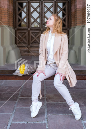 Smiling young woman in white clothes sits on bench with bouquet of flowers on historic building background. Vertical frame. Smiling young woman in white clothes sits on bench with bouquet of flowers on historic building background. Vertical frame. 107964067
