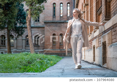 Middle-aged slender woman in white clothes walking near old red brick building. Sad woman alone Middle-aged slender woman in white clothes walking near old red brick building. Sad woman alone 107964080