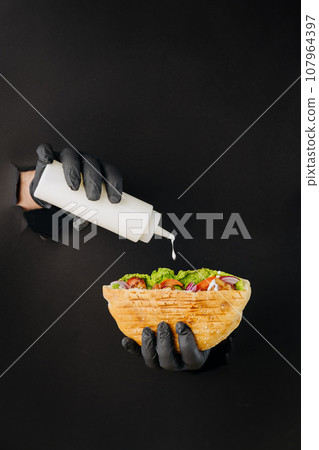 Cooks hand adding sauce to doner kebab through torn holes on black paper background. Street food 107964397