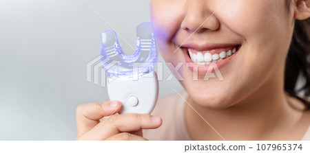 The woman is whitening her teeth with a teeth whitening device by herself in the living room at home. Health and medical concept. 107965374