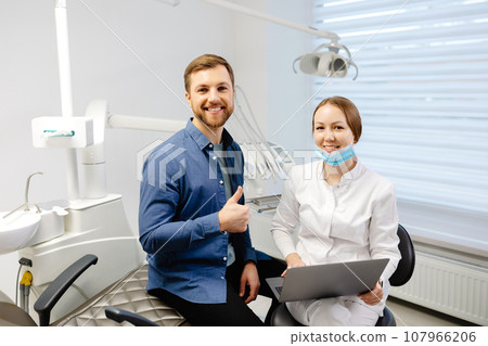 A handsome young man is talking to a female doctor at a dental appointment in a bright, beautiful office. The dentist explains to the patient and shows everything on the laptop 107966206