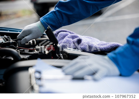 Professional Asian mechanic's hand in blue shirt writing notes on a paper, while working on a car in an auto repair shop. Close-up shot. 107967352