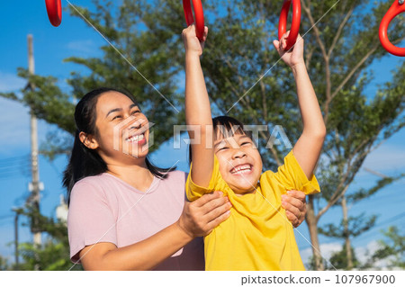 Cute little girl hanging bars in the playground. Happy little Asian child having fun on playground, climbing bars in park. Little girl playing in outdoor playground with mother. 107967900