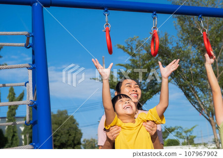Cute little girl hanging bars in the playground. Happy little Asian child having fun on playground, climbing bars in park. Little girl playing in outdoor playground with mother. Cute little girl hanging bars in the playground. Happy little Asian child having fun on playground, climbing bars in park. Little girl playing in outdoor playground with mother. 107967902