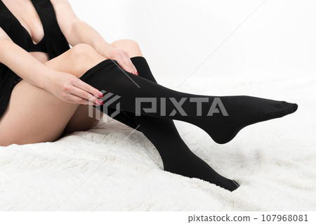 Woman pulling black tight pantyhose over her legs carefully. Crop view of impatient unrecognizable female sitting on bed on white background. Part of series 107968081