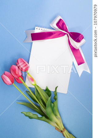 Bouquet of pink tulips and paper for text with lilac bow on blue background. Holiday, Valentine's, International Women's and Mother's Day, March 8, Birthday. Copy space Bouquet of pink tulips and paper for text with lilac bow on blue background. Holiday, Valentine's, International Women's and Mother's Day, March 8, Birthday. Copy space 107970979