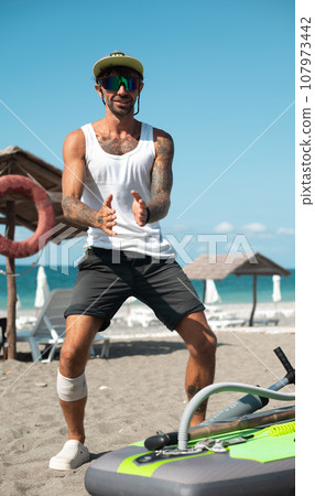 a sporty surfer guy with a pump pumps up an inflatable SUP board for swimming on the seashore a sporty surfer guy with a pump pumps up an inflatable SUP board for swimming on the seashore 107973442
