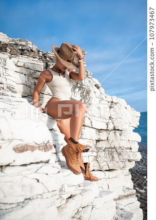 sexy girl in a bodysuit and a cowboy hat and cool boots poses freely sexually on white rocks 107973467